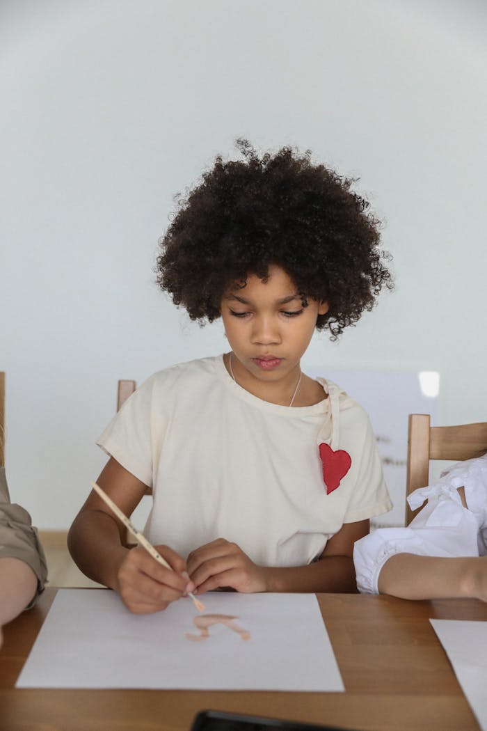 Concentrated talented African American girl with curly hair using paintbrush while drawing with watercolor sitting at table with unrecognizable friends