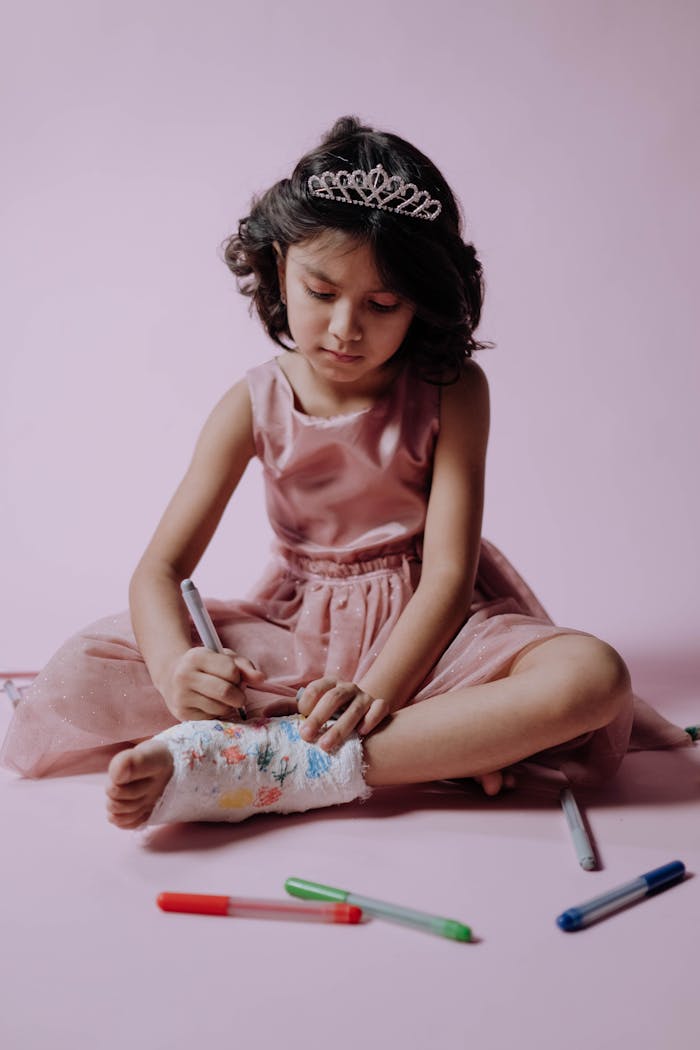 A young girl in a pink dress and tiara decorates her orthopedic cast with markers in a studio setting.